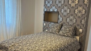 A bedroom with a patterned bedspread, a hanging light fixture, a wall-mounted TV, and patterned wallpaper. - Spacious apartment 15 minutes from Paris (Noisy-le-Sec)