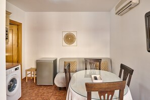Un espacio habitable compacto con lavadora, un refrigerador pequeño, una mesa de vidrio y un sofá con cojines.