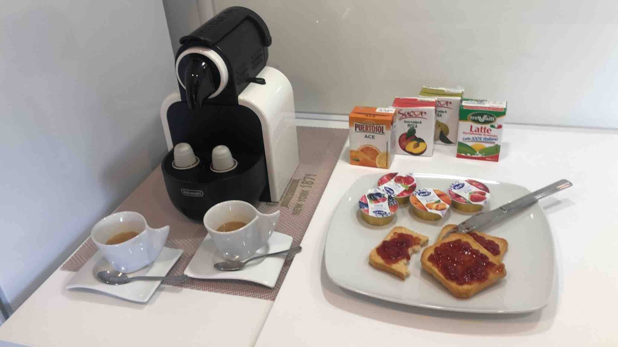 Een ontbijtarrangement met een koffiezetapparaat, twee koffiekopjes, een bord met toast en jam, en diverse ontbijtartikelen zoals ontbijtgranen en vruchtensappen.