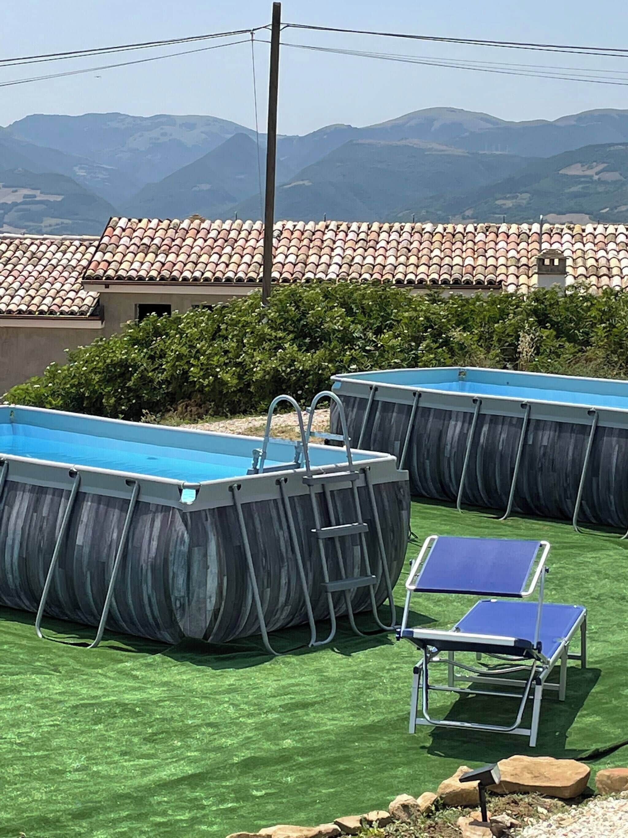 Due piscine fuori terra con scalette, un tavolo da ping pong e una sedia, su un prato sintetico con montagne sullo sfondo.