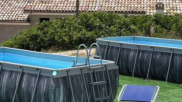 Due piscine fuori terra con scalette, un tavolo da ping pong e una sedia, su un prato sintetico con montagne sullo sfondo.