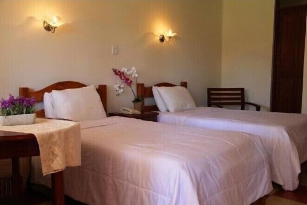 Une chambre d’hôtel avec deux lits simples, une table de chevet en bois avec un téléphone, une petite table sur laquelle est posé un vase de fleurs et des lampes fixées au mur.