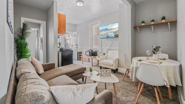 Un salon chaleureux avec un canapé, des fauteuils, une table à manger dressée pour deux personnes et, au fond, un coin cuisine.