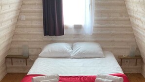 Una cama individual con ropa de cama blanca y una manta roja, dos toallas enrolladas, una ventana con cortinas oscuras y un suelo de madera.