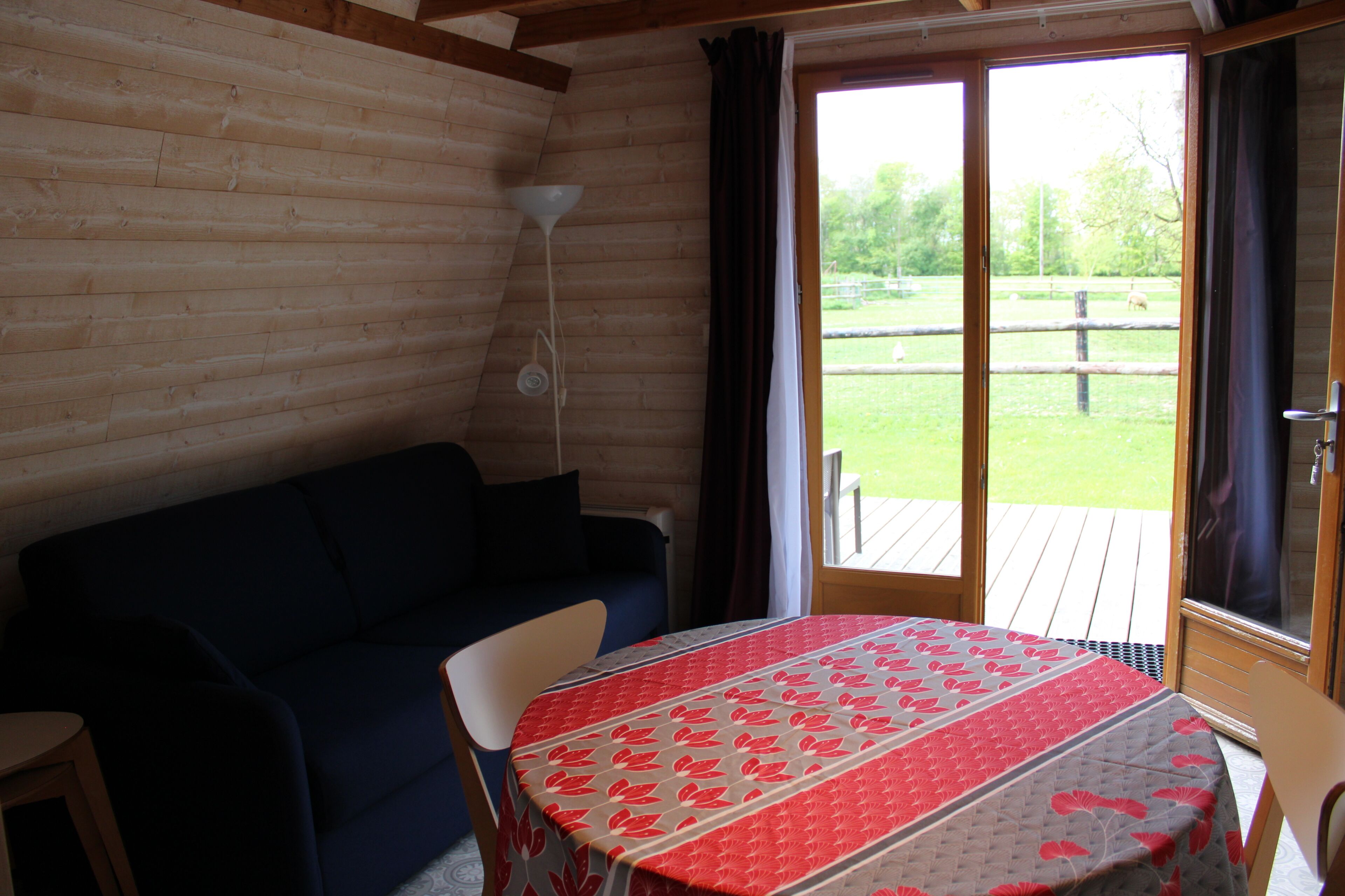 A room with a wooden interior, a round table with a patterned tablecloth, a dark-colored sofa, and a view of a grassy outdoor area through a sliding glass door.