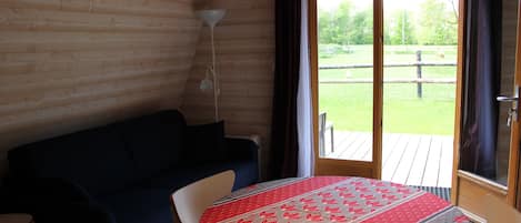 A room with a wooden interior, a round table with a patterned tablecloth, a dark-colored sofa, and a view of a grassy outdoor area through a sliding glass door.