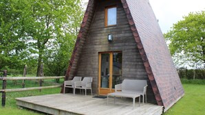 Una estructura de madera tipo cabaña, con puerta de vidrio, terraza de madera con dos sillas y una pequeña mesa.
