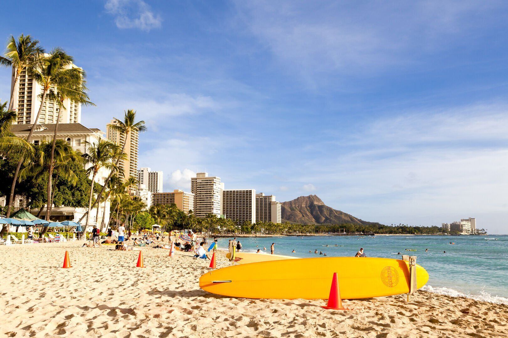A beach with a yellow surfboard, palm trees, and a city skyline in the background.