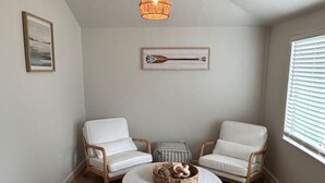 Una habitación con dos sillones de cuero blanco, una mesa redonda de mármol con una cesta tejida, una lámpara colgante y una obra enmarcadas en la pared.