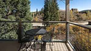 A covered balcony with a table and chairs, overlooking a forested area with autumn foliage and a distant house. - Winter Park Condo w/ Fireplace, Centrally Located (Winter Park)
