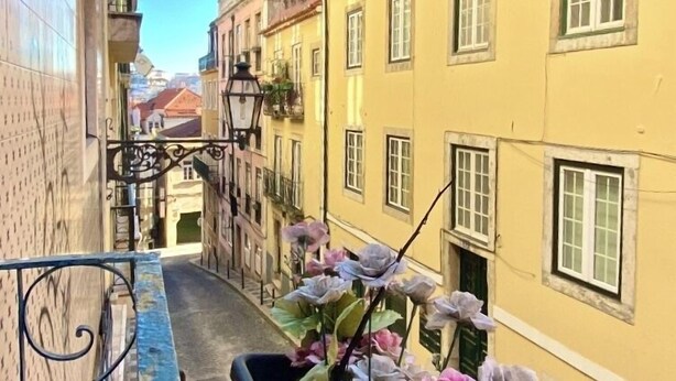Une rue étroite bordée d’immeubles jaunes, avec une rambarde métallique bleue et un jardinier où sont plantées des fleurs roses.