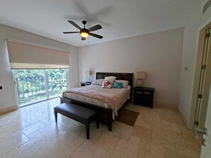 A bedroom with a bed, bedside tables, a bench, a ceiling fan, and a balcony with a view of greenery. - Luxury Near the Sea Buenaventura | Beach, Pool, Golf and Resort Life in Río Hato (Buenaventura)
