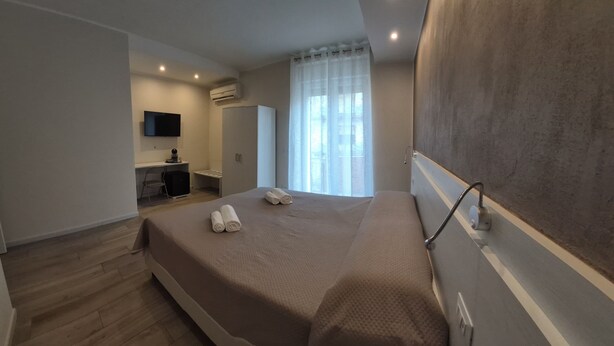 A hotel room with a bed, a desk with a chair, a TV mounted on the wall, and a window with curtains.