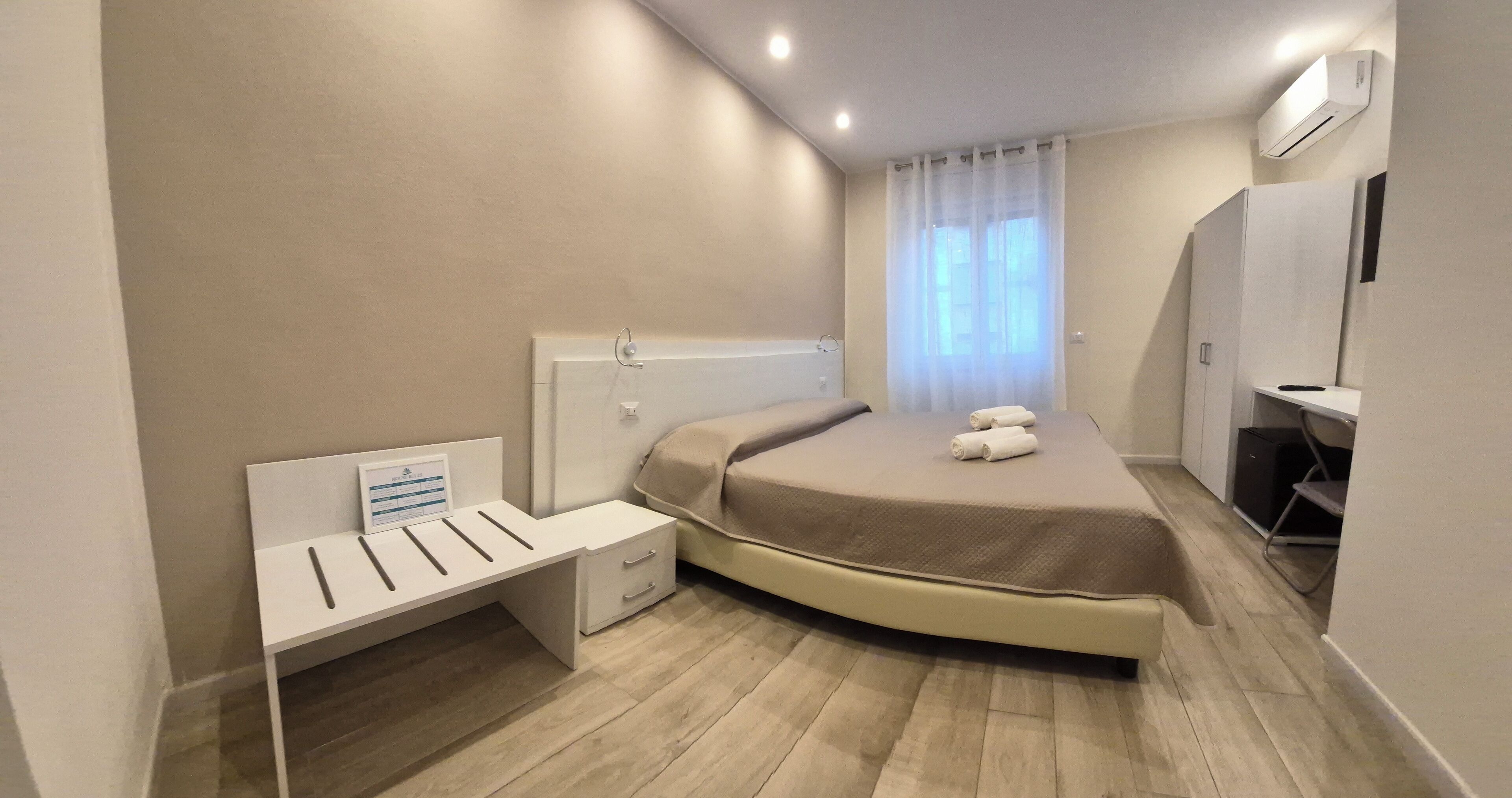 A modern bedroom with a curved bed, a small bedside table with drawers, a desk, and a window with sheer curtains.