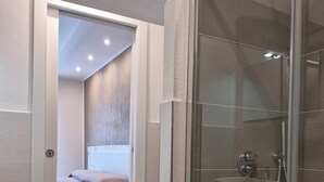 A modern bathroom with a glass shower enclosure, a visible toilet, and a bed in the adjacent room.