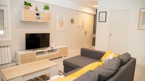 A modern living room with a grey sofa, a wooden coffee table, and a TV on a white stand. There are two potted plants on a shelf above the TV. - Cool Vialia (Vigo)