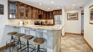 A kitchen with wooden cabinets, a marble countertop, and a built-in bar with stools. - VIEWS! Corner Unit Next to River - Steps to Ski Slopes & Downtown Festivities (Breckenridge)