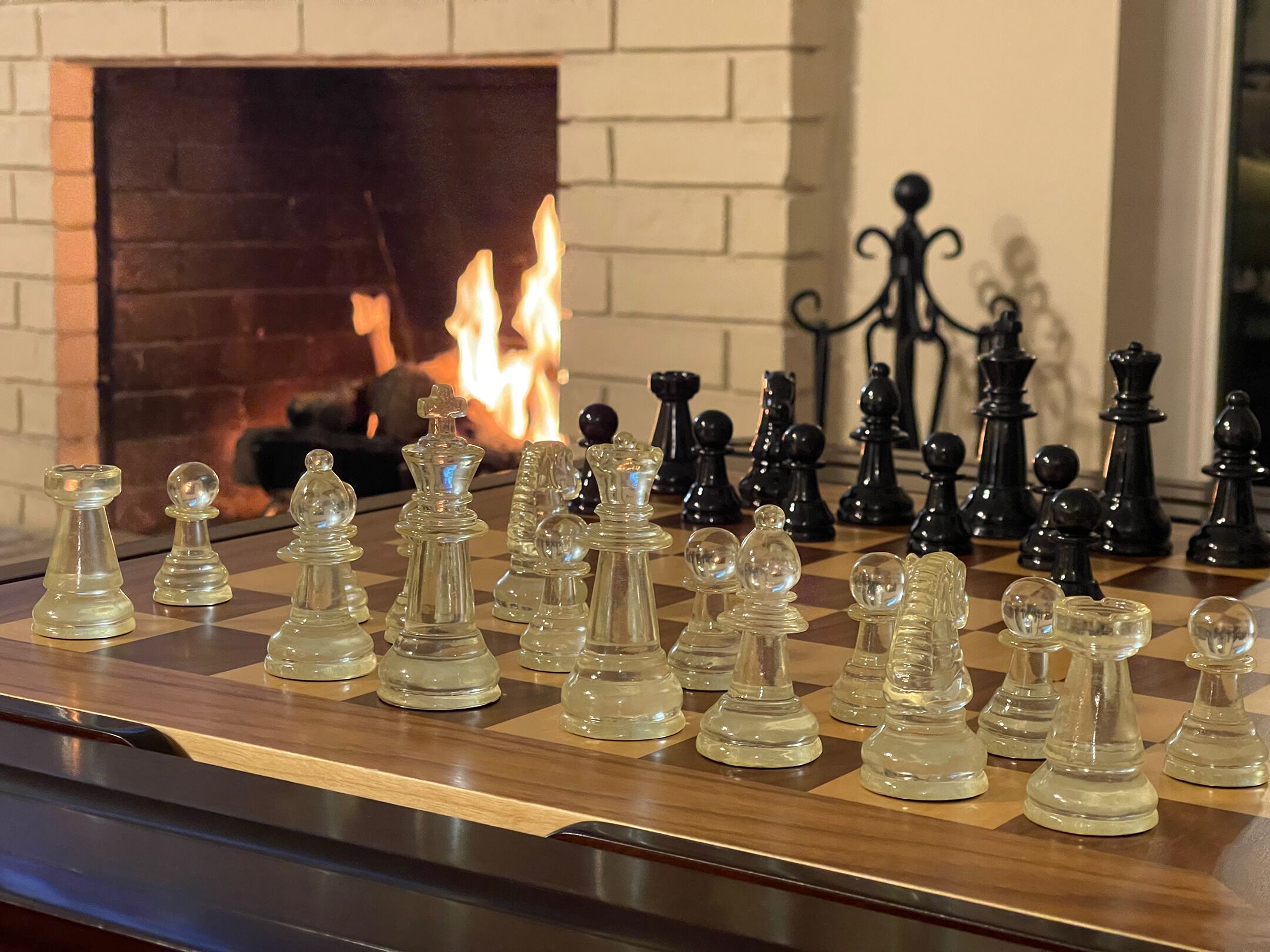 A chessboard with glass and black pieces in front of a fireplace.