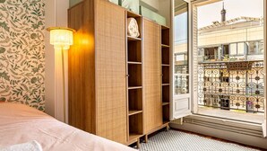 Een slaapkamer met een bed, een houten kledingkast, een vloerlamp en een raam met uitzicht op een gebouw en een metalen hek.