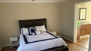 Een slaapkamer met een bed, nachtkastjes, een houten hoofdbord, een plafondventilator en een zichtbare badkamer door een open deur.