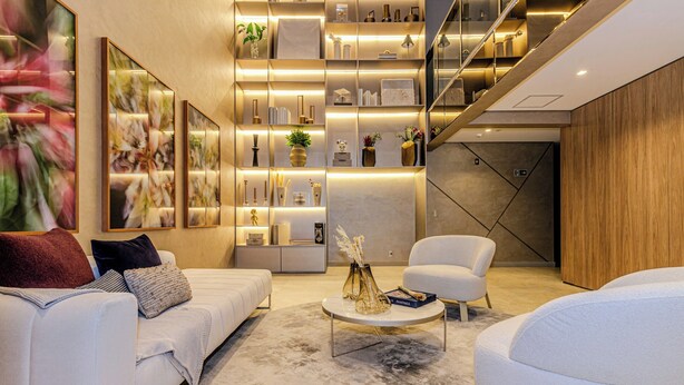 Een moderne woonkamer met een bank, fauteuil en salontafel, met een marmeren vloer en een boekenplank met decoratieve objecten.