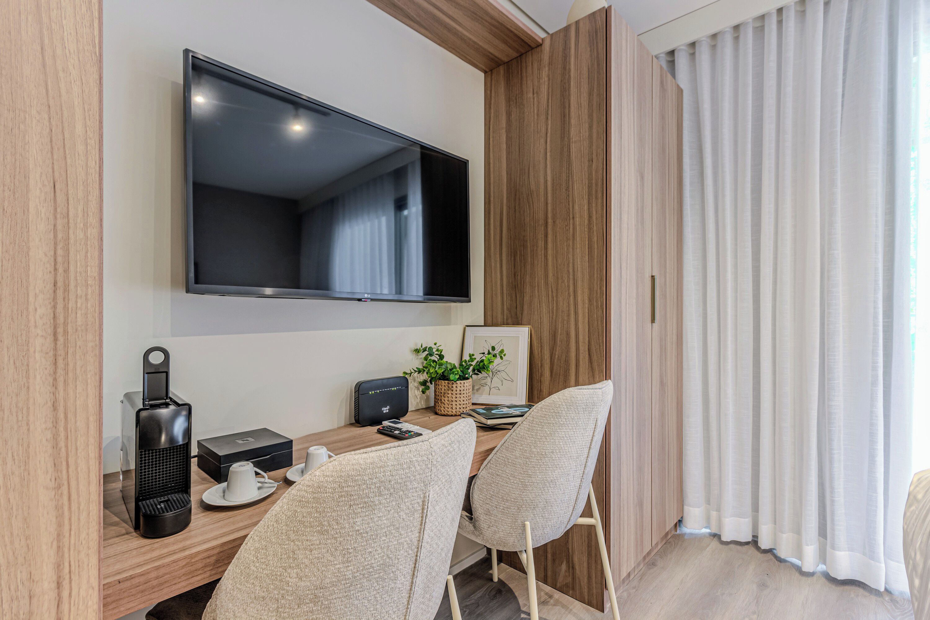 A modern room with a flat-screen TV mounted on the wall, a wooden desk with a coffee maker, a plant, and a framed picture.