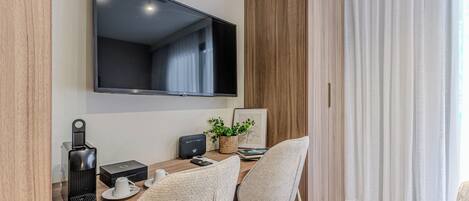 A modern room with a flat-screen TV mounted on the wall, a wooden desk with a coffee maker, a plant, and a framed picture.