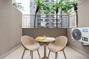 A balcony with two wicker chairs, a small table with a basket and a book, and an air conditioning unit. - Charlie Le Jardin Pinheiros - Soft Opening (São Paulo)