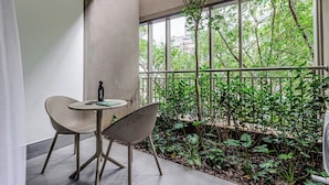 Een balkon met twee rieten stoelen, een klein rond tafeltje en een fles. Er is uitzicht op groen en bomen door een metalen hekwerk.