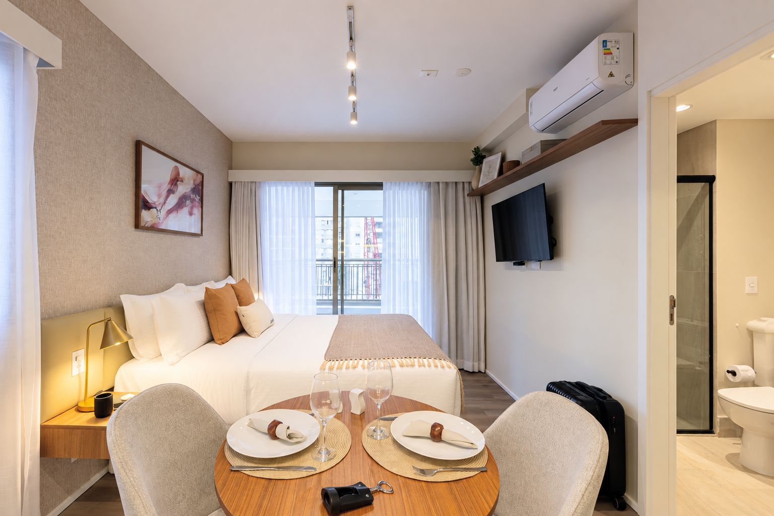 Habitación de hotel con cama, mesita de noche, lámpara, televisión y zona de comedor con mesa para dos.