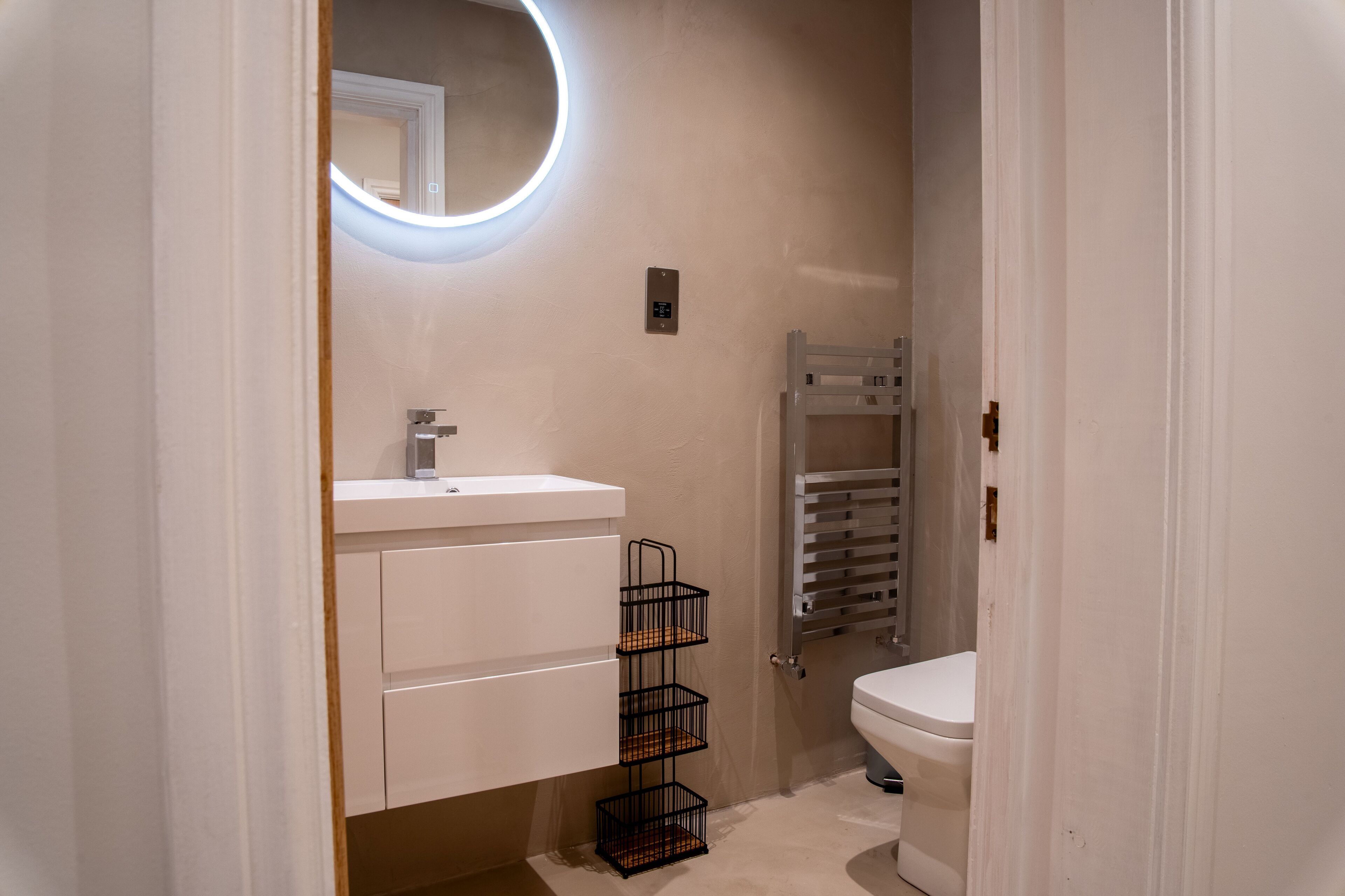 Une salle de bain moderne avec un lavabo blanc, un miroir doté d’un éclairage circulaire, un porte-serviettes et des toilettes.