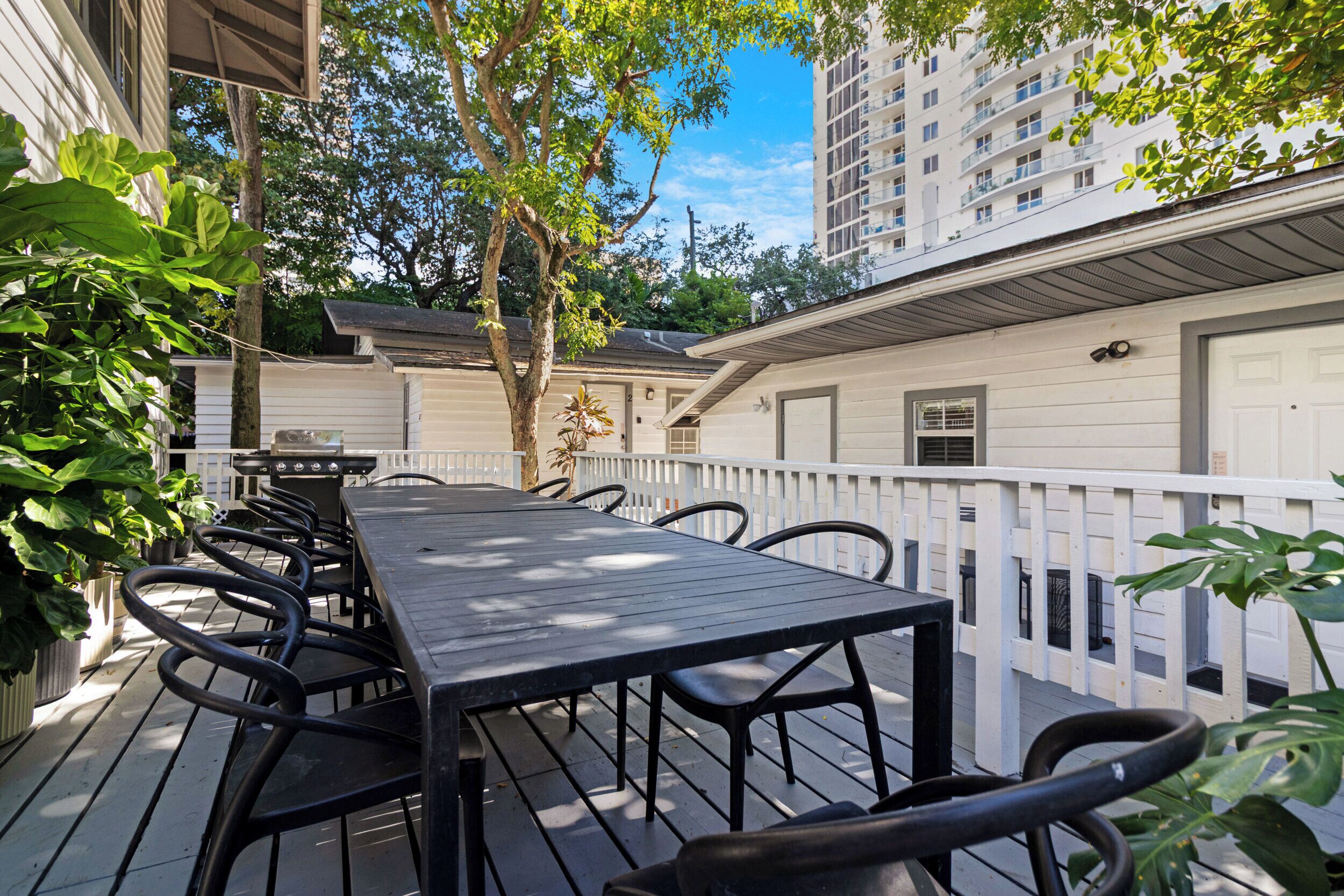 A deck with a long table and chairs, surrounded by a white railing, with a building in the background.