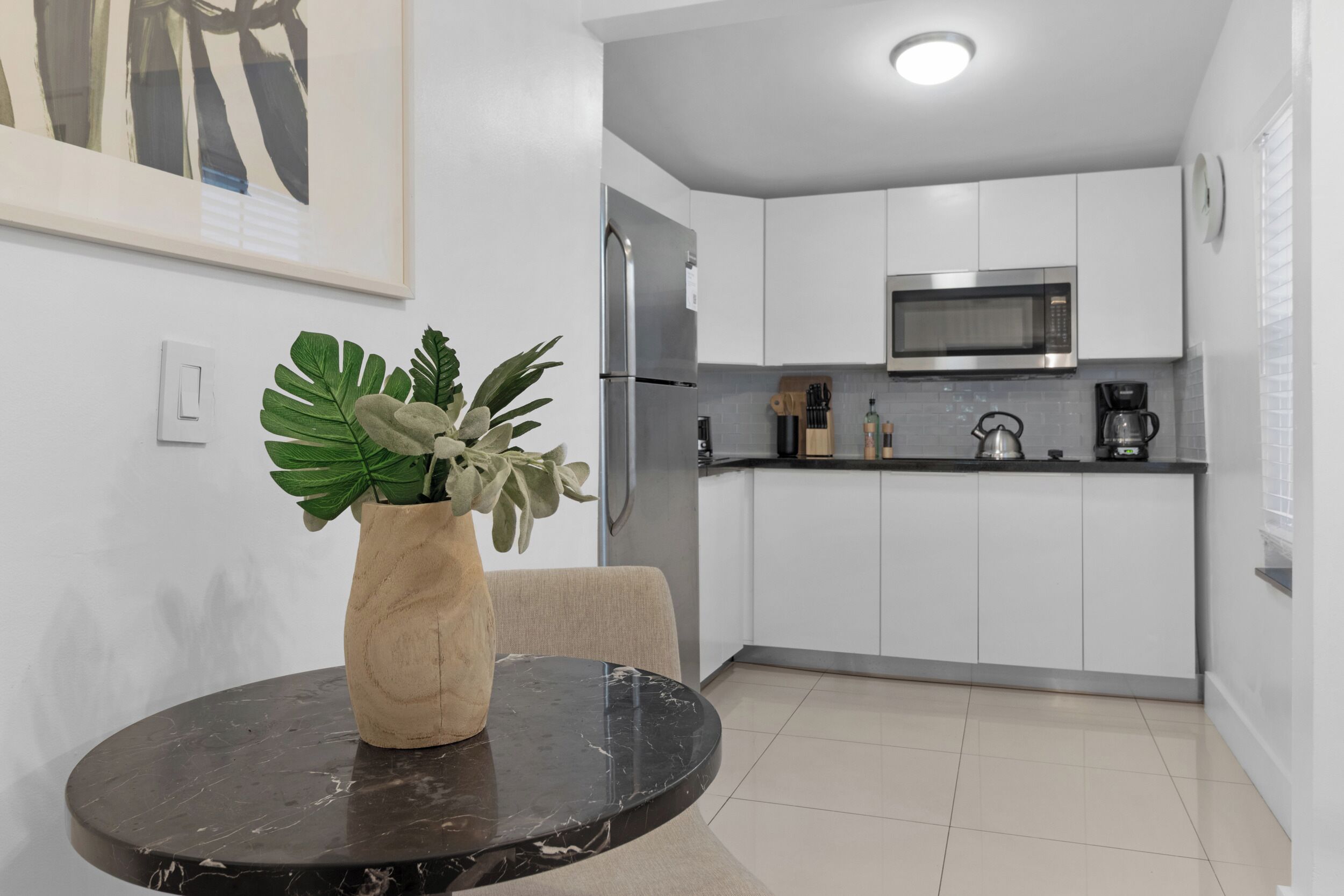 A modern kitchen with white cabinets, a stainless steel refrigerator, a microwave, and a coffee maker. There is a round table with a vase of green plants.