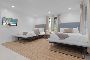 A modern bedroom with two beds, a large window with curtains, a small bedside table, and a wall-mounted TV displaying a beach scene. - 7BD Downtown Oasis Pool  (Miami)