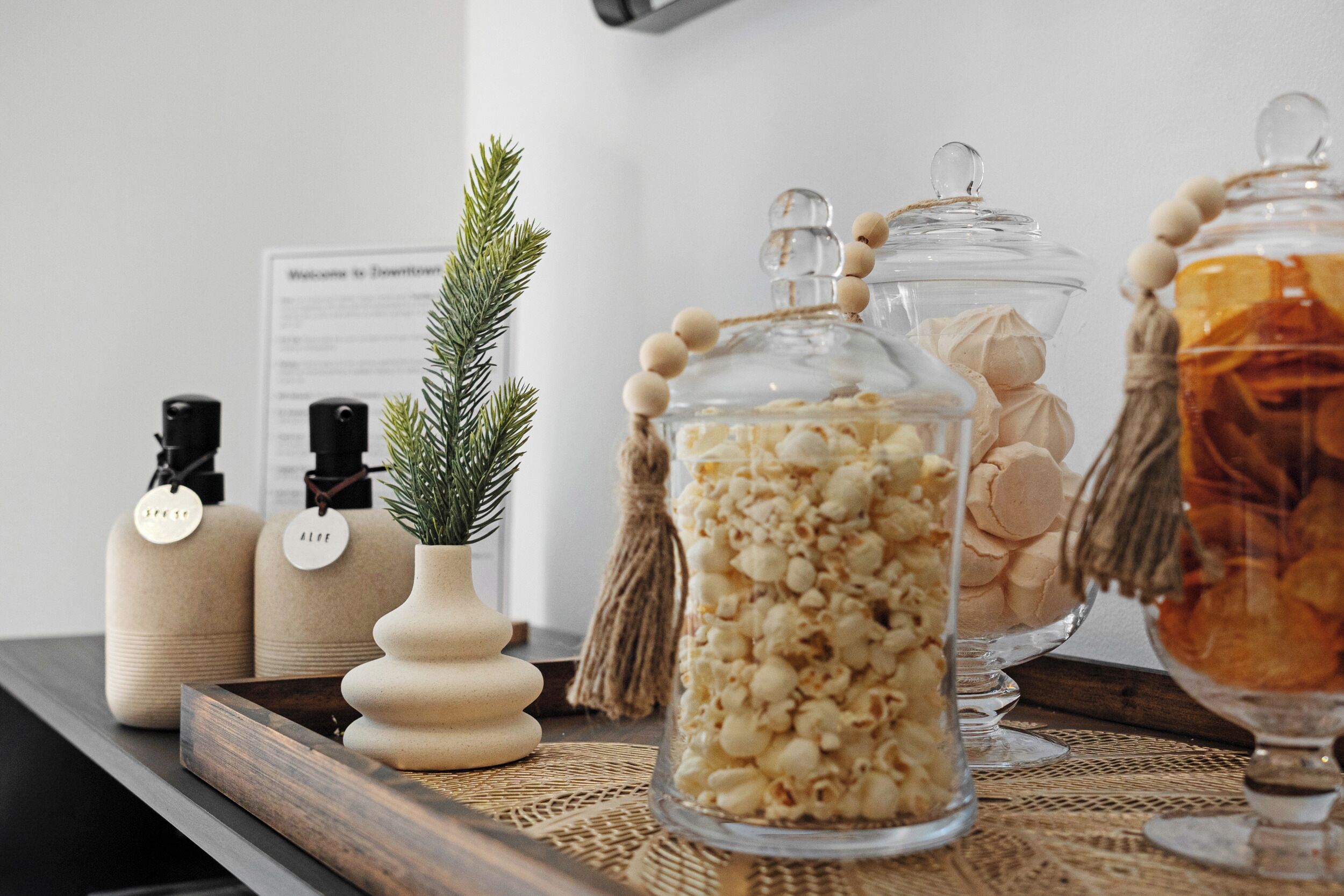 A wooden tray with glass jars containing popcorn and other snacks, a small potted plant, and two bottles labeled "SPICE" and "ALOE."