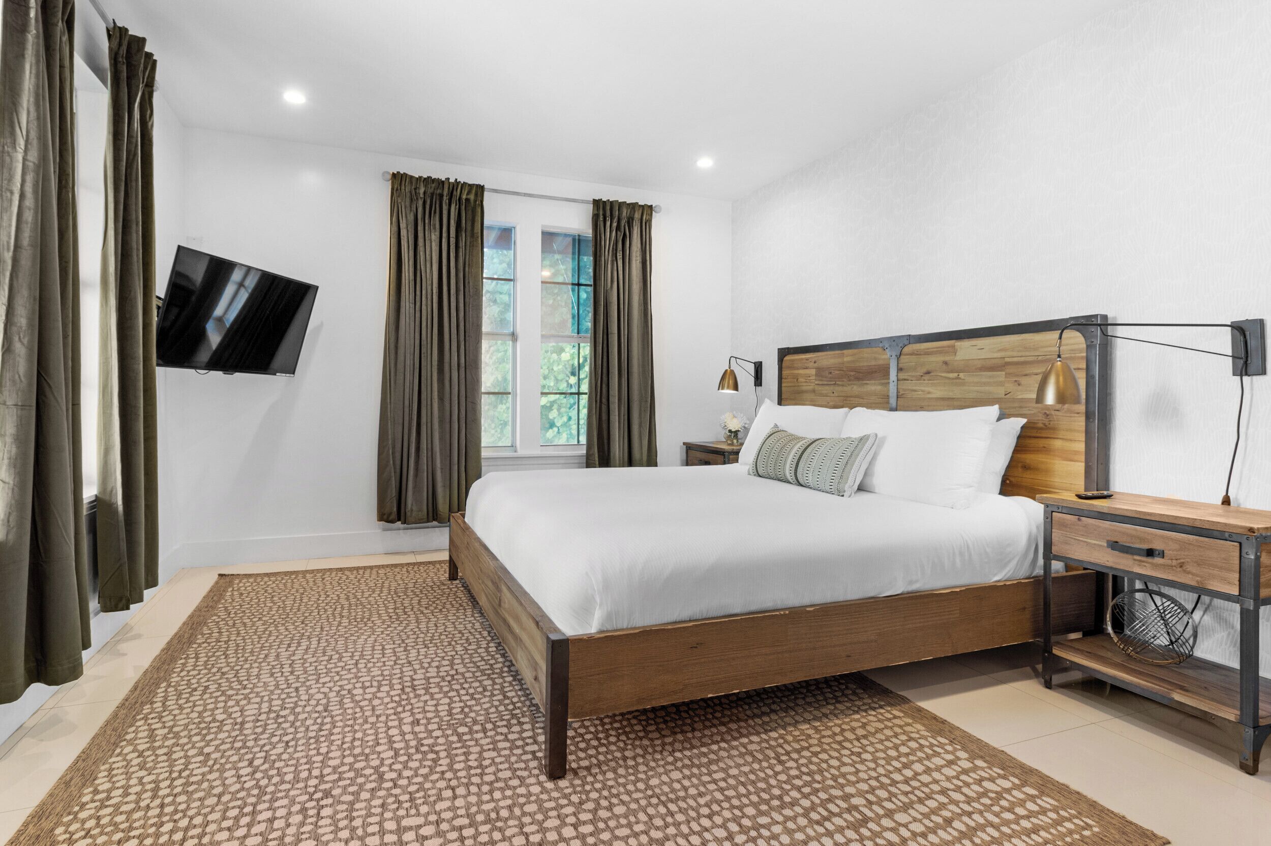 A bedroom with a wooden bed, a TV mounted on the wall, a bedside table with a lamp, and a window with curtains.