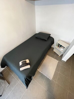 A small, single-bed room with a dark-colored bedspread, a pillow, and a bedside table with a book. The room has a tiled floor and a white wall. - Private room in Colonia Roma Norte. (Ciudad de México)