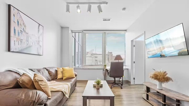 A modern living room with a leather sofa, a coffee table, a TV, and a large window with blinds. - DT · Fast Wifi · Pet Friendly · Bidet Sprayer (Cleveland)