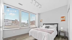 A bedroom with a bed, a nightstand, a window with blinds, and a view of buildings outside. - DT · Fast Wifi · Pet Friendly · Bidet Sprayer (Cleveland)