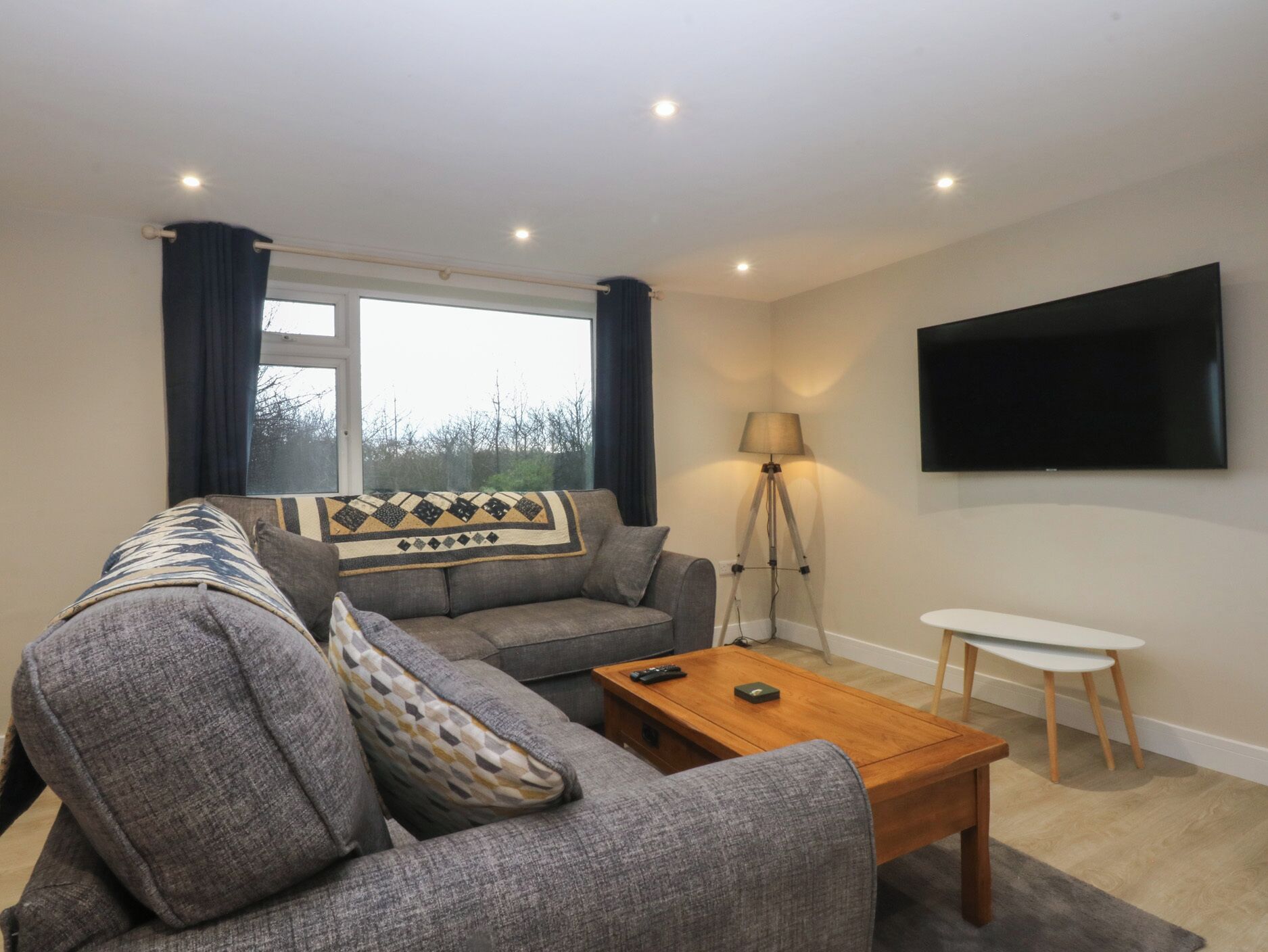 Een moderne woonkamer met een grijze bank, een houten salontafel, een vloerlamp en een flatscreen televisie die aan de muur is bevestigd.