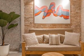 Una habitación acogedora con una pared de ladrillo, un sofá color beige con cojines estampados, y un cuadro abstracto grande sobre el sofá.