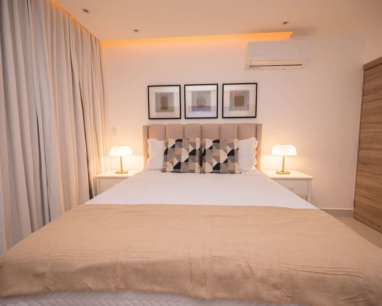 A neatly made bed with a geometric patterned pillow, two bedside lamps, and a wall-mounted air conditioner.