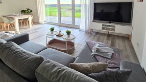 Ein modernes Wohnzimmer mit einer großen, grauen Couch, einem Flachbildfernseher und einem Essbereich mit Tisch und Stühlen.