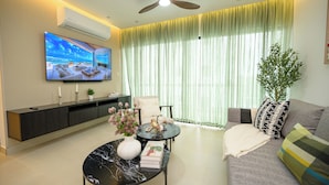 Uma sala de estar moderna com televisão de tela plana, ventilador de teto, sofá, mesa de centro e uma grande janela com cortinas finas.