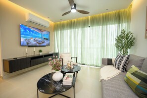 A modern living room with a flat-screen TV, a ceiling fan, a sofa, a coffee table, and a large window with sheer curtains. - Accommodates 15 Guests | 2 Full Apartments in the Same Building (Santiago de los Caballeros)