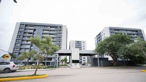 Un edificio moderno de varios pisos con una entrada central, rodeado de árboles y un estacionamiento.