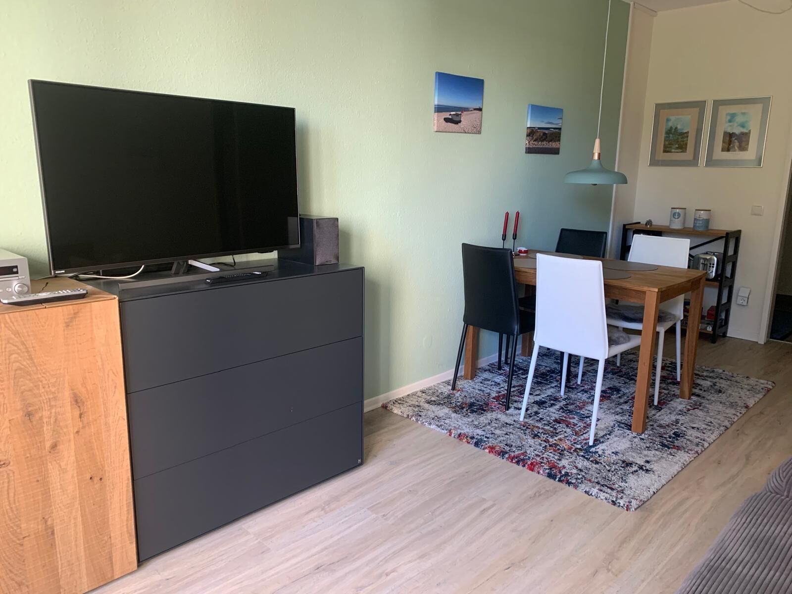 Un saló modern amb un televisor de pantalla plana sobre un moble negre, una zona de menjador amb una taula i cadires de fusta, i una catifa amb dibuixos.