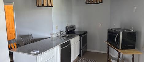 A compact kitchen with a granite countertop, stainless steel appliances, and a wooden hanging light fixture.
