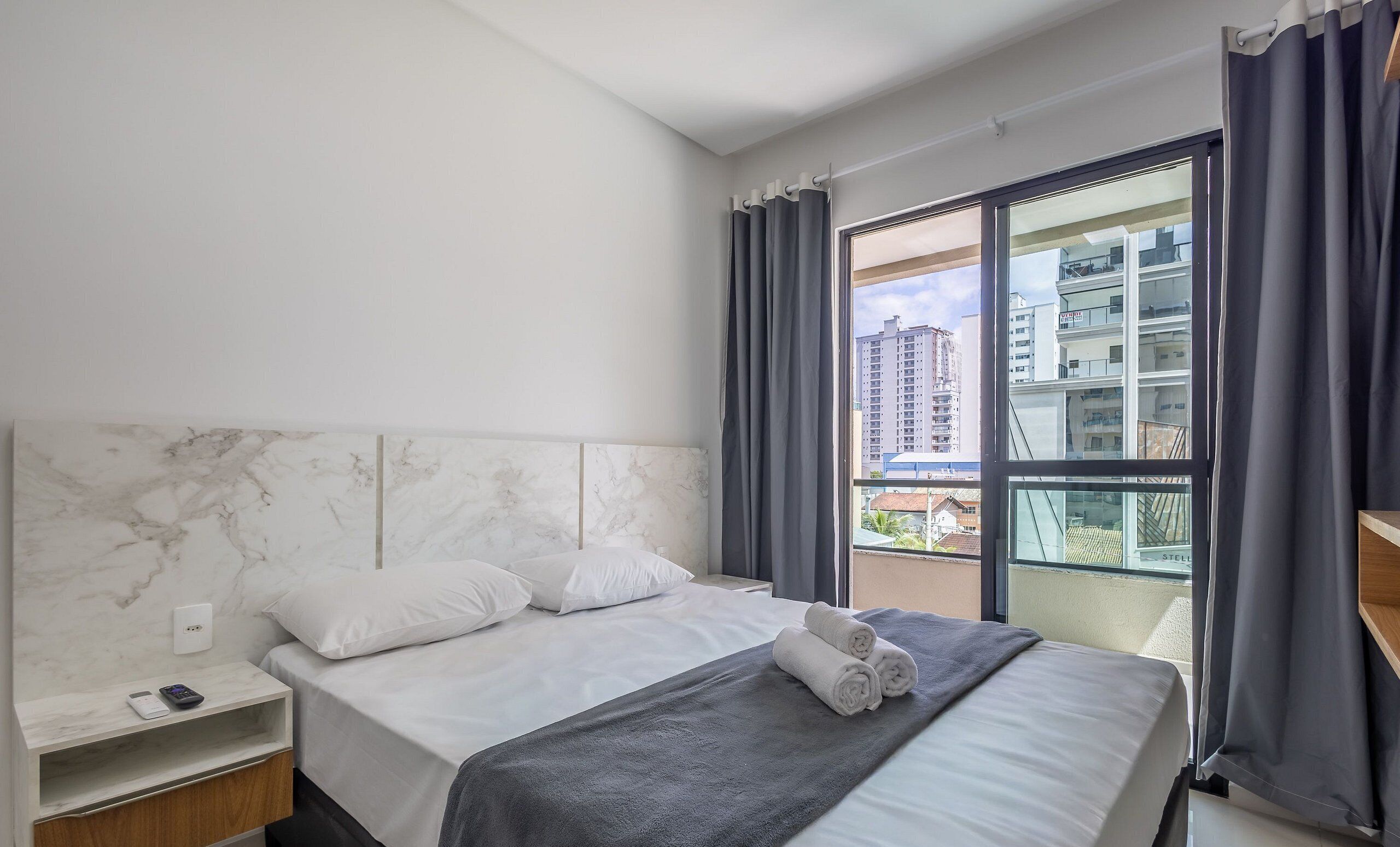 A hotel room with a marble headboard, a bed with white linens, a grey blanket, and a view of buildings through a large window.