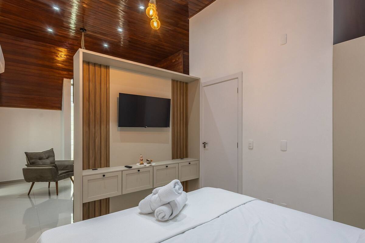 Ein modernes Hotelzimmer mit Holzboden, einem flachbildfernseher an der Wand, einer Minibar, einem Bett mit zusammengerolltem Handtuch und einem Sessel.